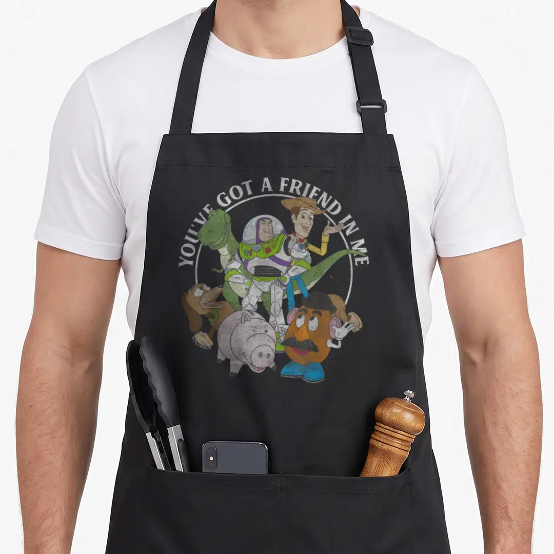 Cute shot friends cooking apron - Cartoon group of toys with Buzz Lightyear and text YOU'VE GOT A FRIEND IN ME arched — shown on a black cotton canvas apron against a white background