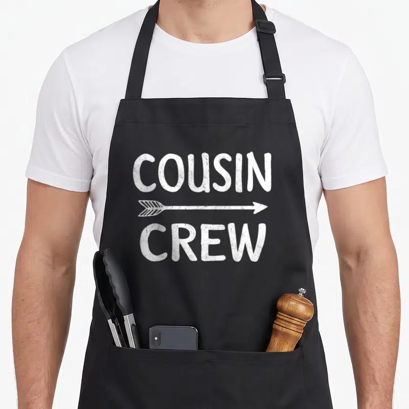 Cousin family crew apron - White distressed COUSIN CREW text with horizontal arrow on black bib — shown on a black cotton canvas apron against a white background