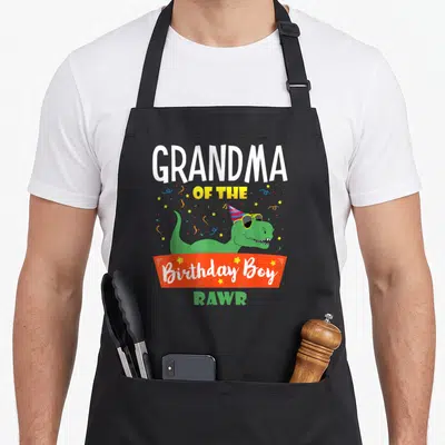 Family grandma in apron - Green dinosaur with party hat and Grandpa of the Birthday Boy text on orange banner — shown on a black cotton canvas apron against a white background