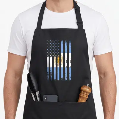 Argentinian american flag apron - Distressed flag merging blue white stripes and yellow sun with white stars on black — shown on a black cotton canvas apron against a white background