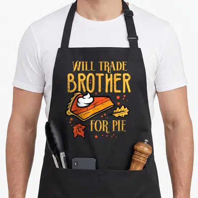 Trade thanksgiving apron - Will Trade Brother For Pie text with pie slice, cream, and autumn leaves — shown on a black cotton canvas apron against a white background
