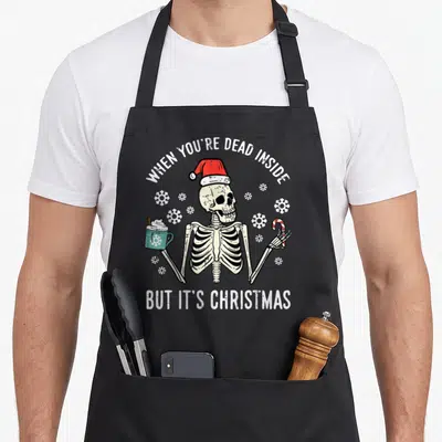 Inside coffee apron - Cartoon skeleton in red hat holding teal mug and candy cane with white snowflakes — shown on a black cotton canvas apron against a white background