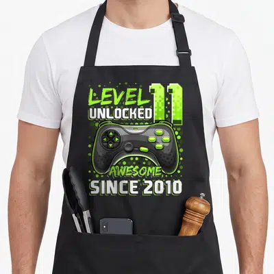 Unlocked 11th birthday video game apron - Black game controller with lime buttons and pixel 11 text reading Level 11 — shown on a black cotton canvas apron against a white background