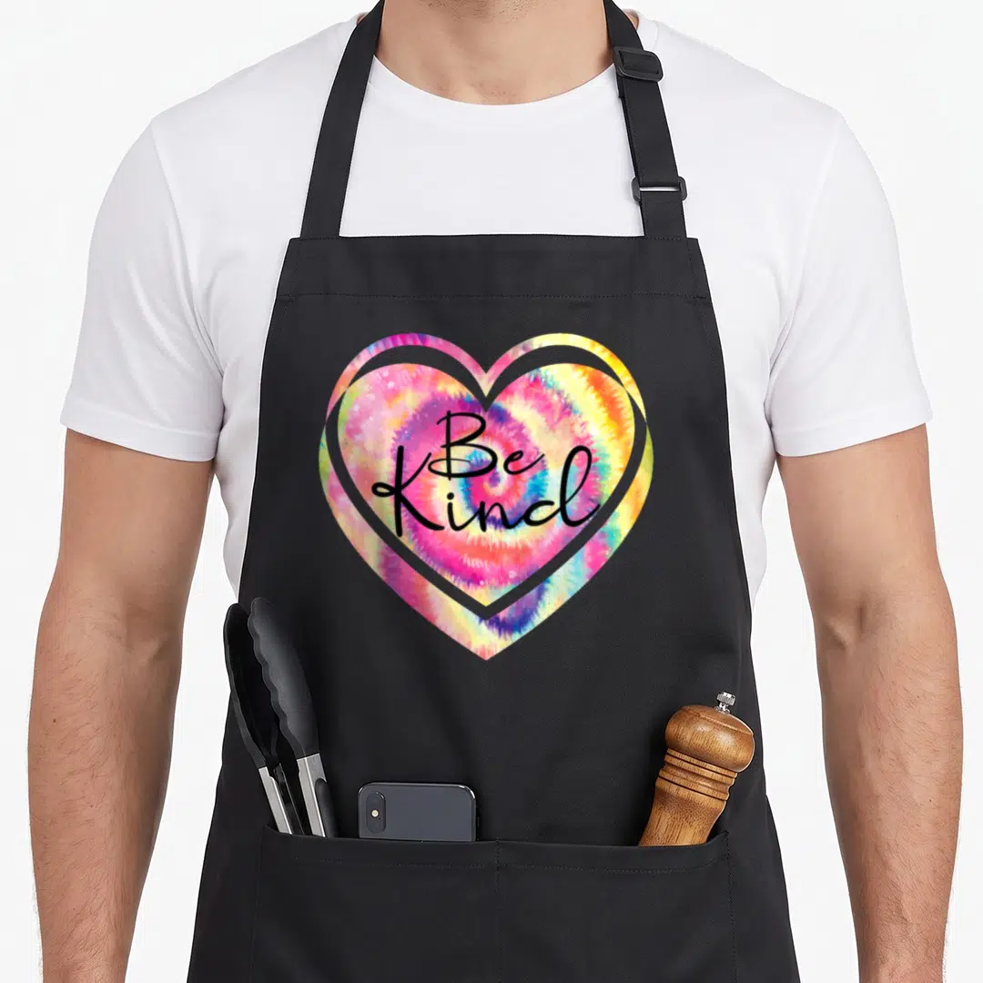 Inspirational hippie apron - Black Be Kind script overlaid on a multi-colored tie-dye heart design — shown on a black cotton canvas apron against a white background