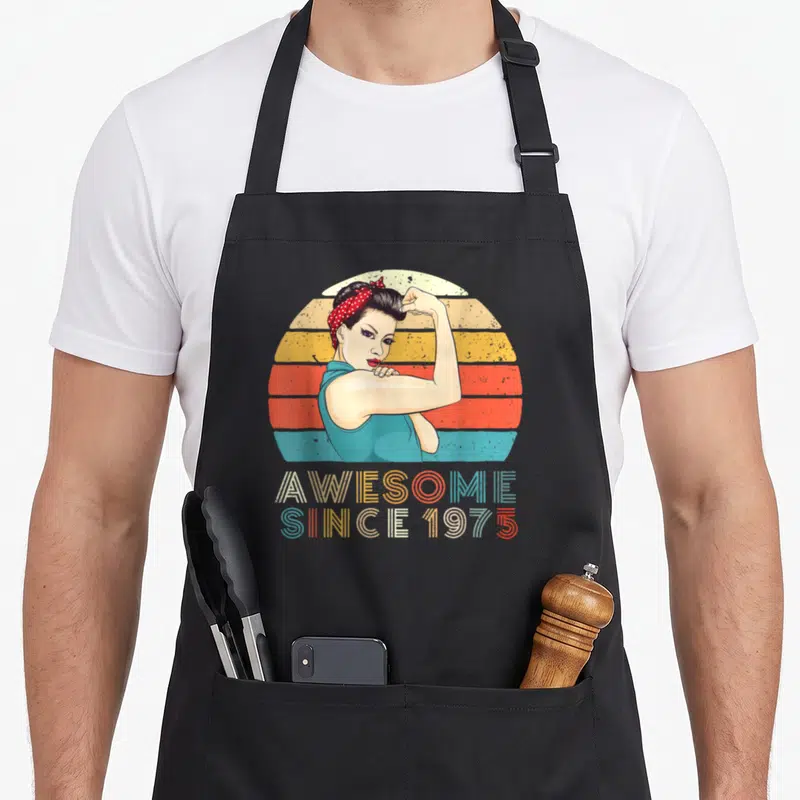 Strong birth birthday apron - Retro woman flexing arm in front of teal orange yellow striped sun with AWESOME SINCE — shown on a black cotton canvas apron against a white background