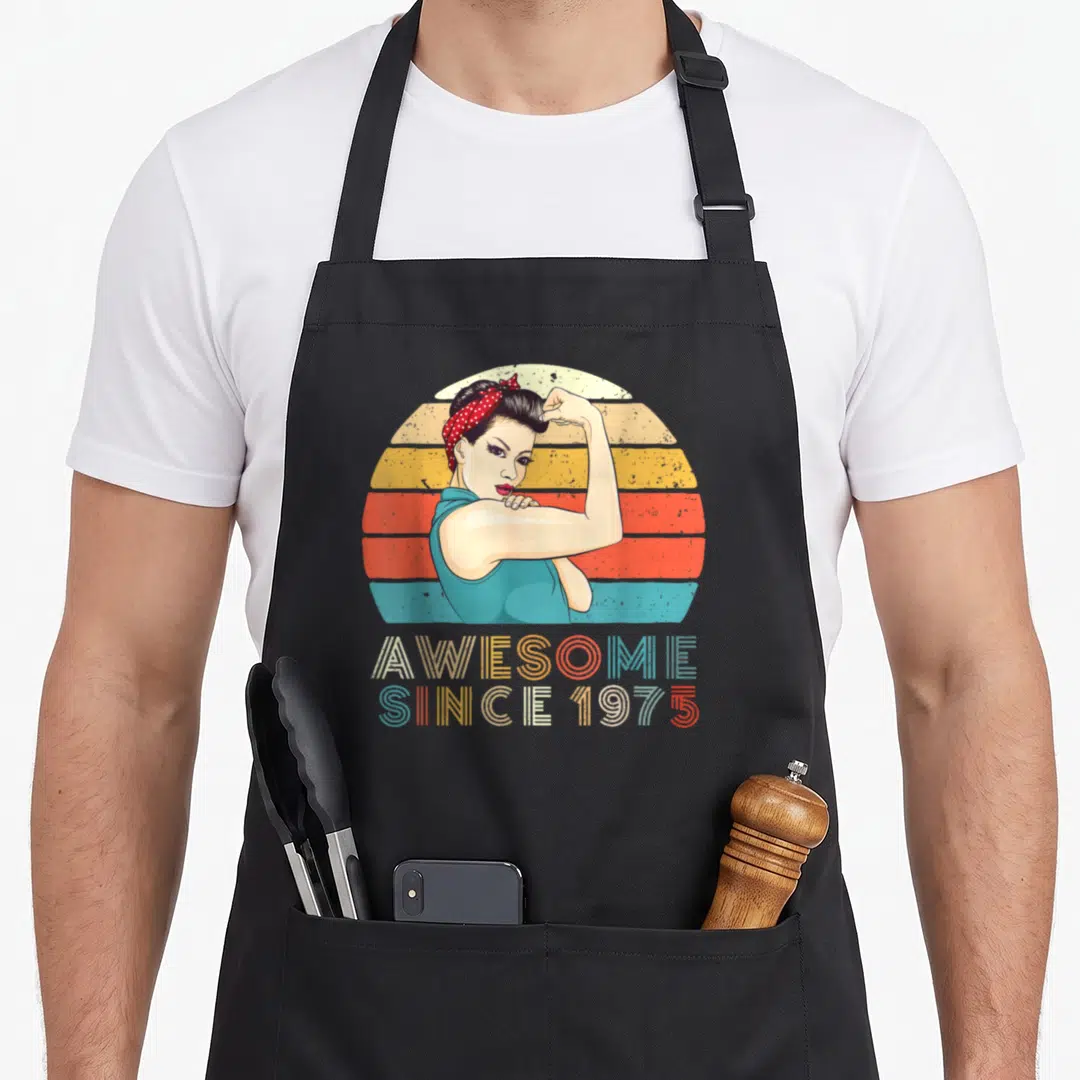 Strong birth birthday apron - Retro woman flexing arm in front of teal orange yellow striped sun with AWESOME SINCE — shown on a black cotton canvas apron against a white background