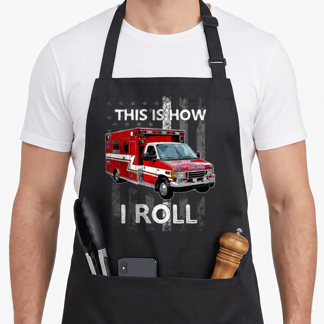 Roll ambulance girl apron - Red and white ambulance on distressed flag with text THIS IS HOW I ROLL — shown on a black cotton canvas apron against a white background