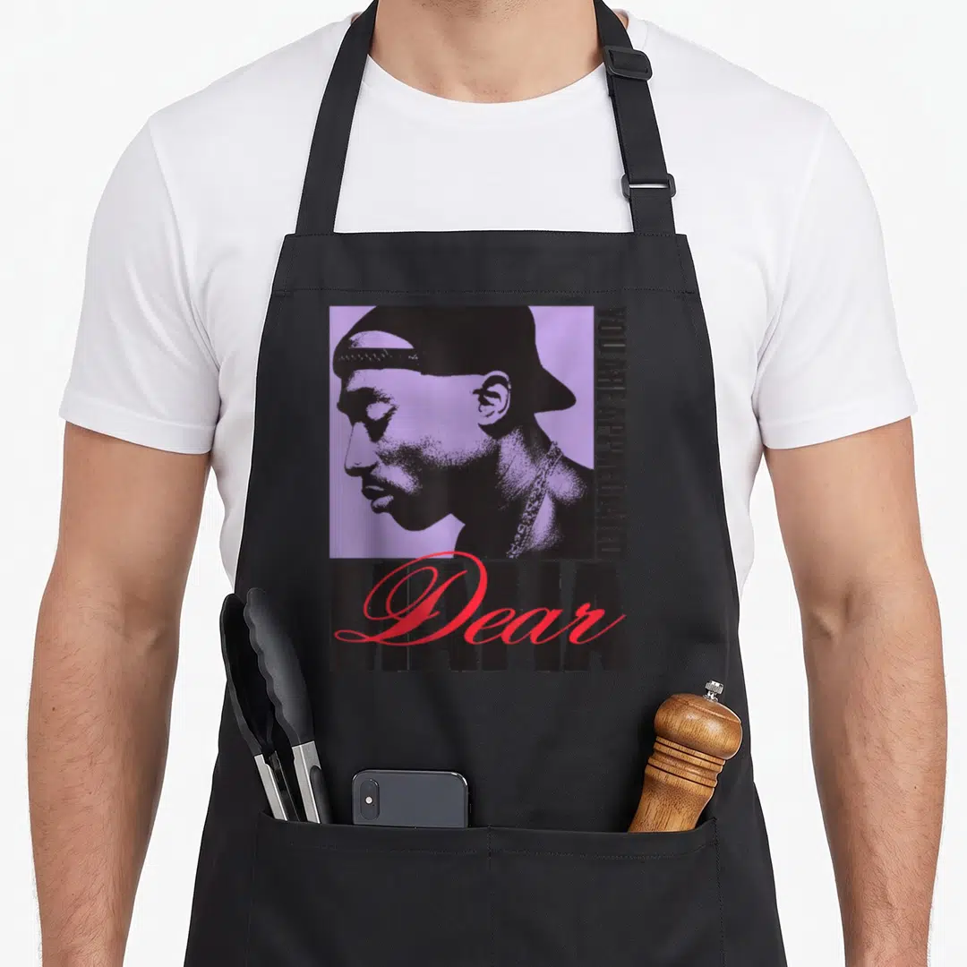 Tupac mama apron - Black man profile on purple square with vertical appreciated text and red Dear Mama script — shown on a black cotton canvas apron against a white background