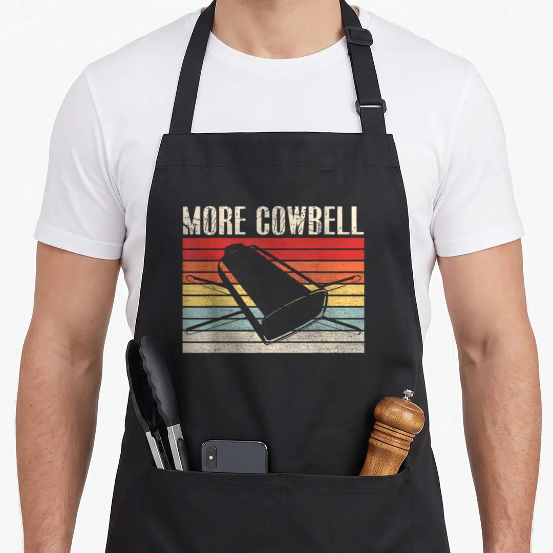 Cowbell sarcastic apron - MORE COWBELL text above grey cowbell silhouette on red orange yellow blue retro stripes — shown on a black cotton canvas apron against a white background