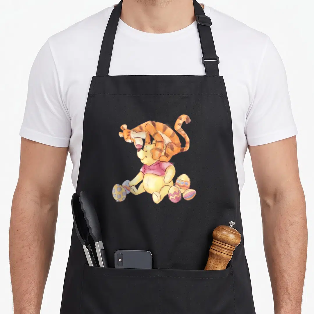 Tigger egg apron - Watercolor Winnie the Pooh sitting with Tigger draped over him among decorated Easter eggs — shown on a black cotton canvas apron against a white background
