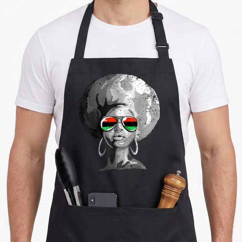 Queen face apron - Woman with large afro and hoop earrings wearing sunglasses with red black and green flag reflection — shown on a black cotton canvas apron against a white background