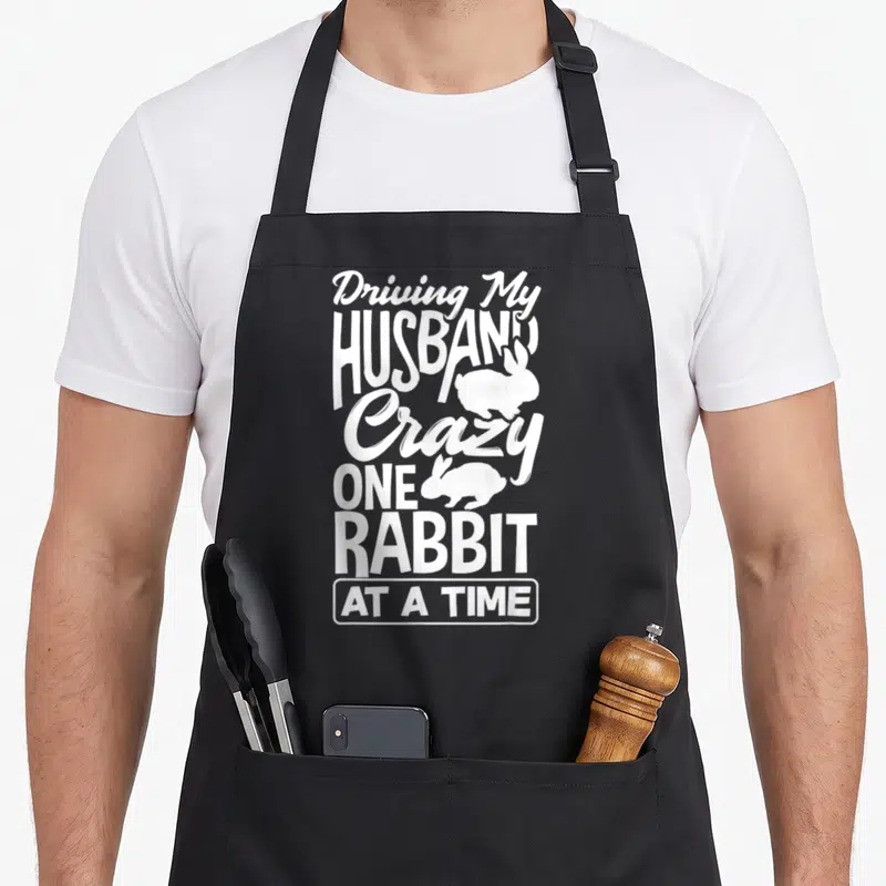 Husband rabbit driving aprons - White stacked text reading Driving My Husband Crazy One Rabbit At A Time with two — shown on a black cotton canvas apron against a white background