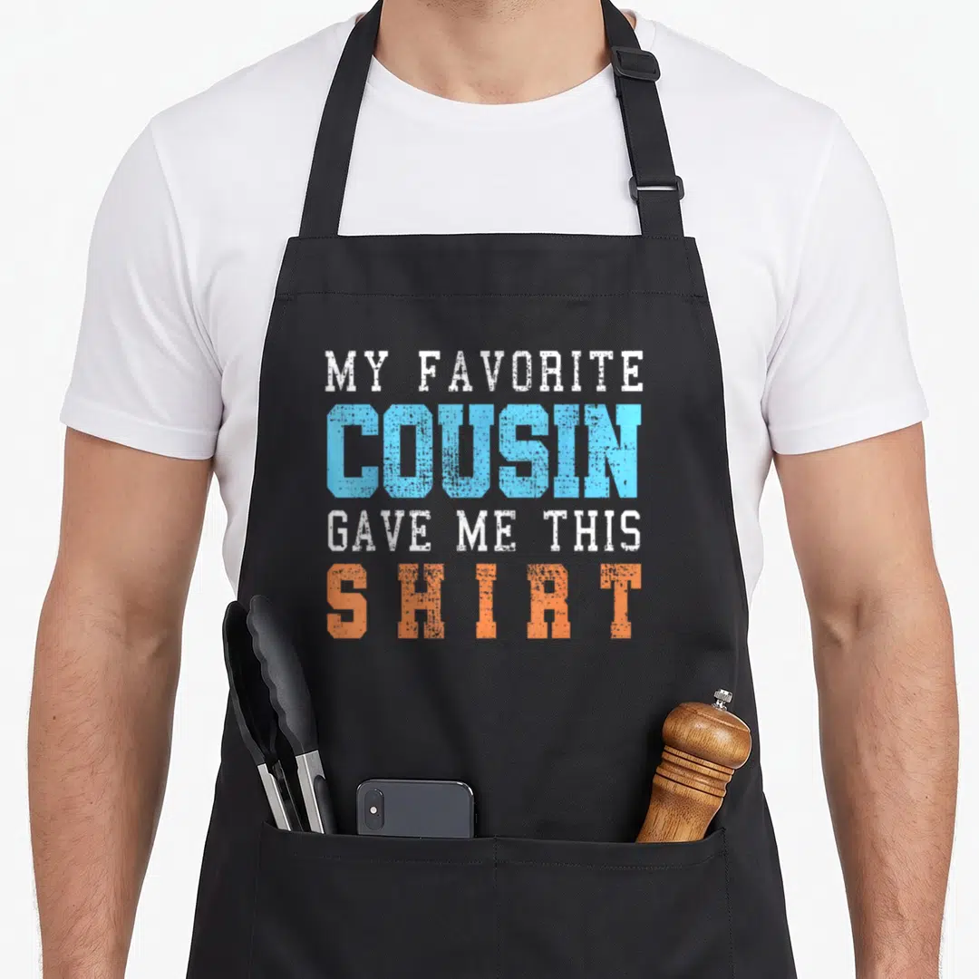 Funny cousin apron - Stacked white blue and orange text reading My Favorite Cousin Gave Me This Shirt — shown on a black cotton canvas apron against a white background