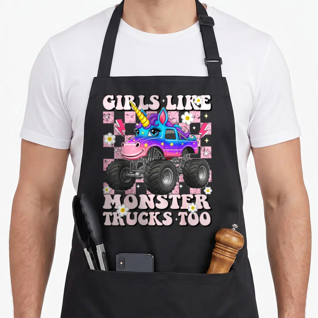 Funny unicorn truck apron - GIRLS LIKE MONSTER TRUCKS TOO text with pink purple blue unicorn truck on checkerboard — shown on a black cotton canvas apron against a white background