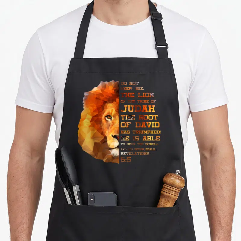 Christ lions apron - Geometric lion head in orange and brown next to stacked Revelations 5:5 text — shown on a black cotton canvas apron against a white background