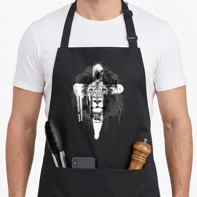 Funny scripture christian apron - Lion head with white cross and paint splatters featuring Revelation 5:5 text — shown on a black cotton canvas apron against a white background