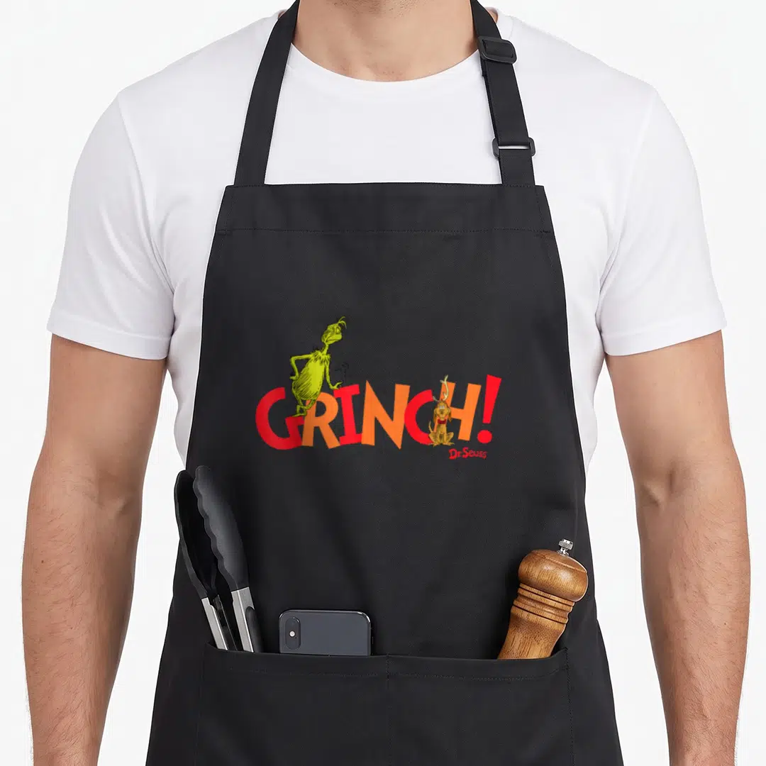 Reindeer holiday apron - The Grinch standing on GRINCH text with Max dog sitting in letters — shown on a black cotton canvas apron against a white background