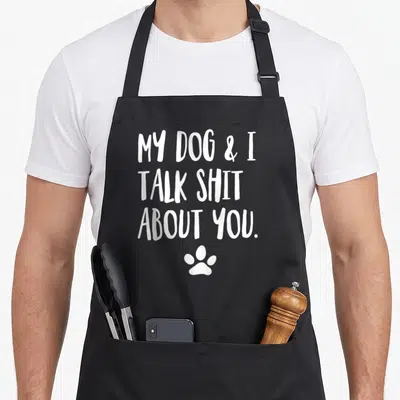 Offensive dog apron - Three lines of white text reading MY DOG & I TALK SHIT ABOUT YOU above a single paw print — shown on a black cotton canvas apron against a white background