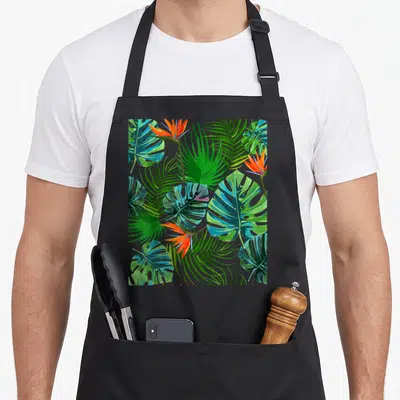 Paradise the floral apron - Cartoon tropical leaves and flowers in green, orange, blue, and teal — shown on a black cotton canvas apron against a white background