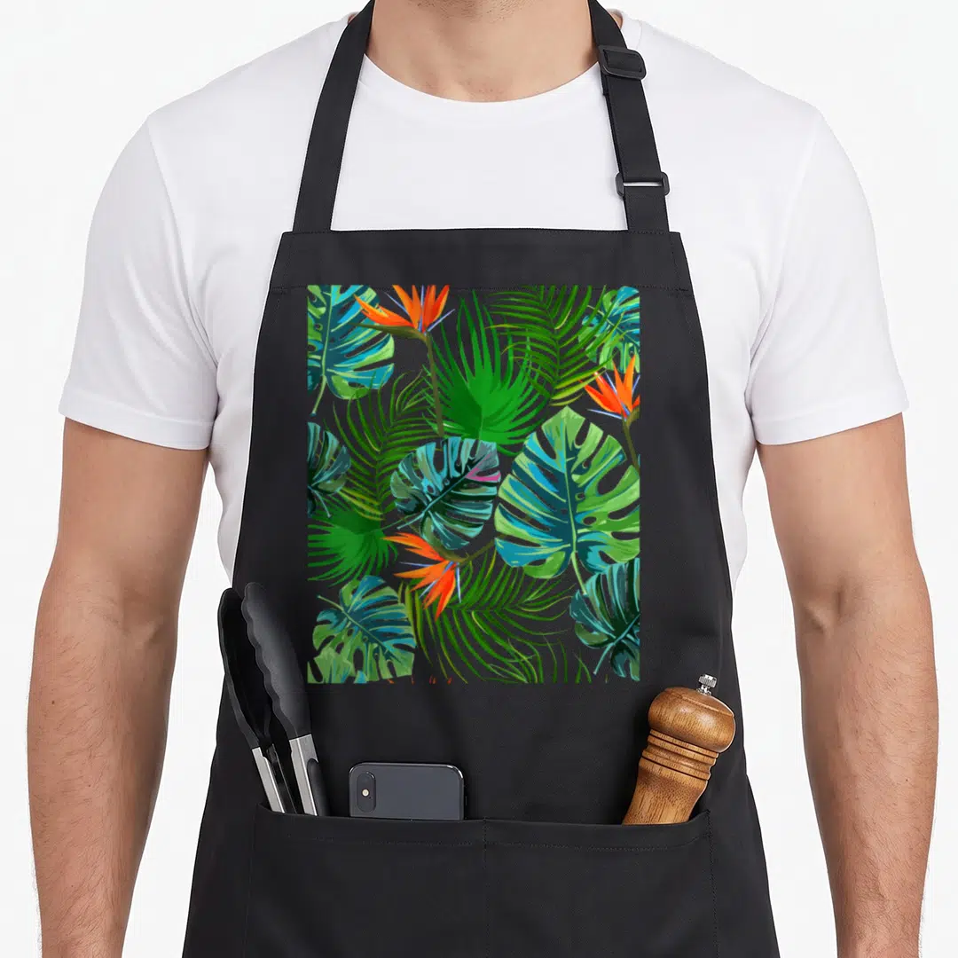 Paradise the floral apron - Cartoon tropical leaves and flowers in green, orange, blue, and teal — shown on a black cotton canvas apron against a white background