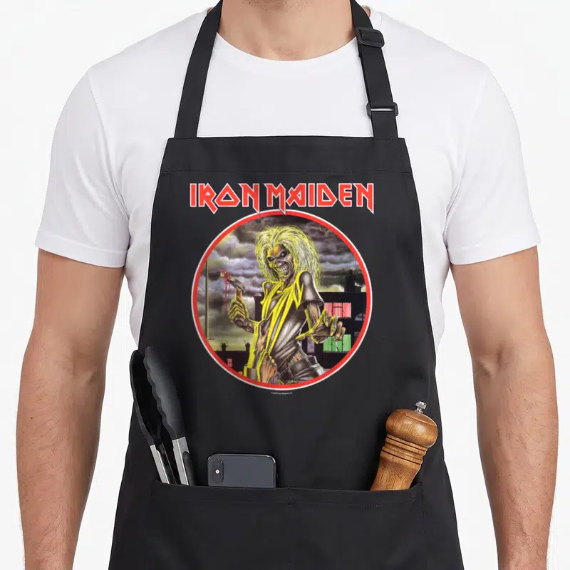 Funny camping apron - IRON MAIDEN text above Eddie standing in a circle with stormy skyline background — shown on a black cotton canvas apron against a white background