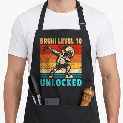 Dabbing bruh 10th birthday apron - Retro boy dabbing holding game controller over red orange yellow cyan grey stripes — shown on a black cotton canvas apron against a white background