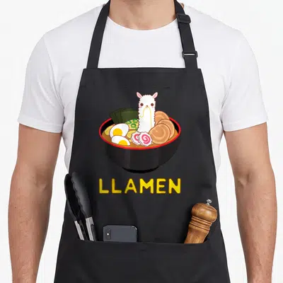 Llamen anime apron - White llama in black ramen bowl with pink fish cakes and yellow LLAMEN text — shown on a black cotton canvas apron against a white background
