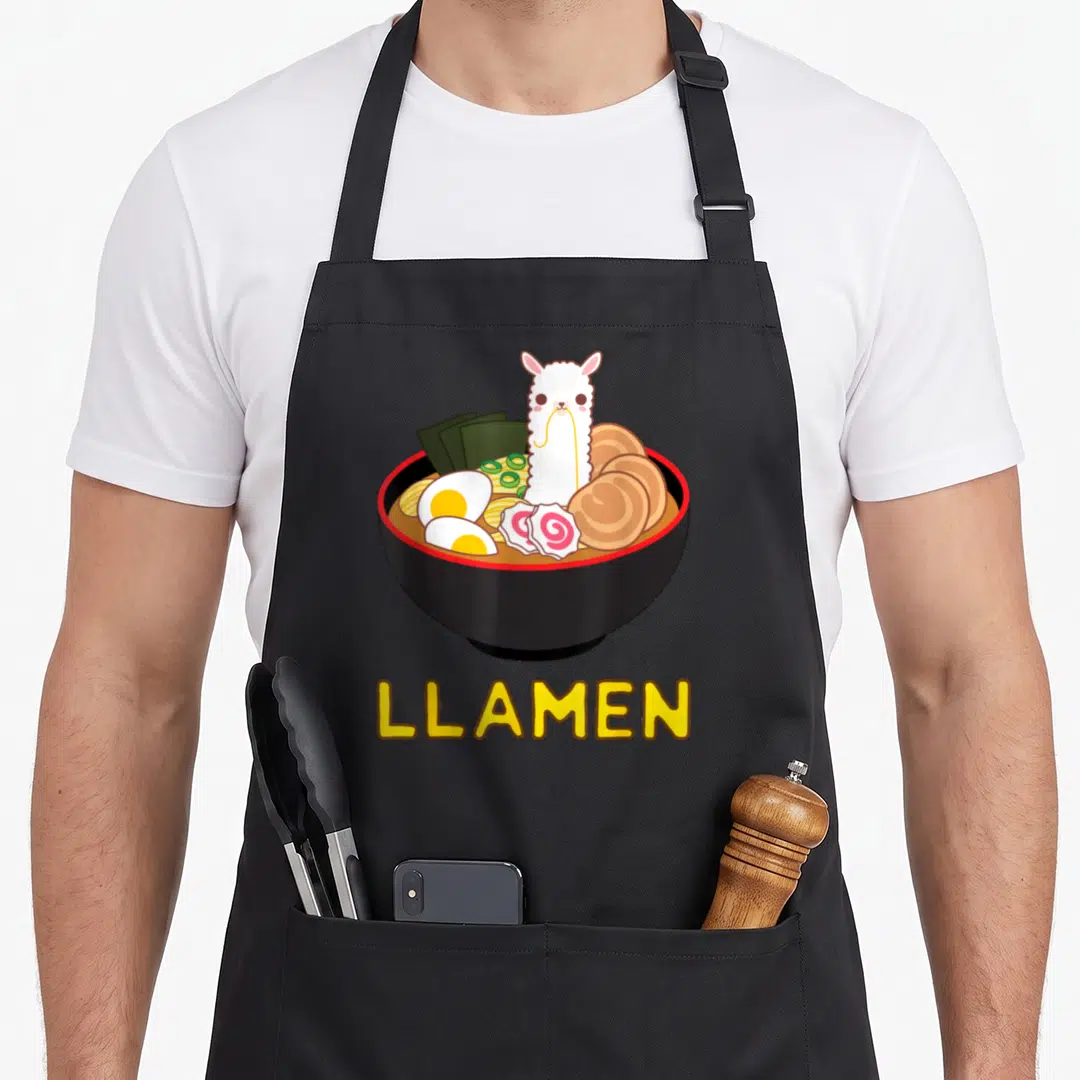 Llamen anime apron - White llama in black ramen bowl with pink fish cakes and yellow LLAMEN text — shown on a black cotton canvas apron against a white background