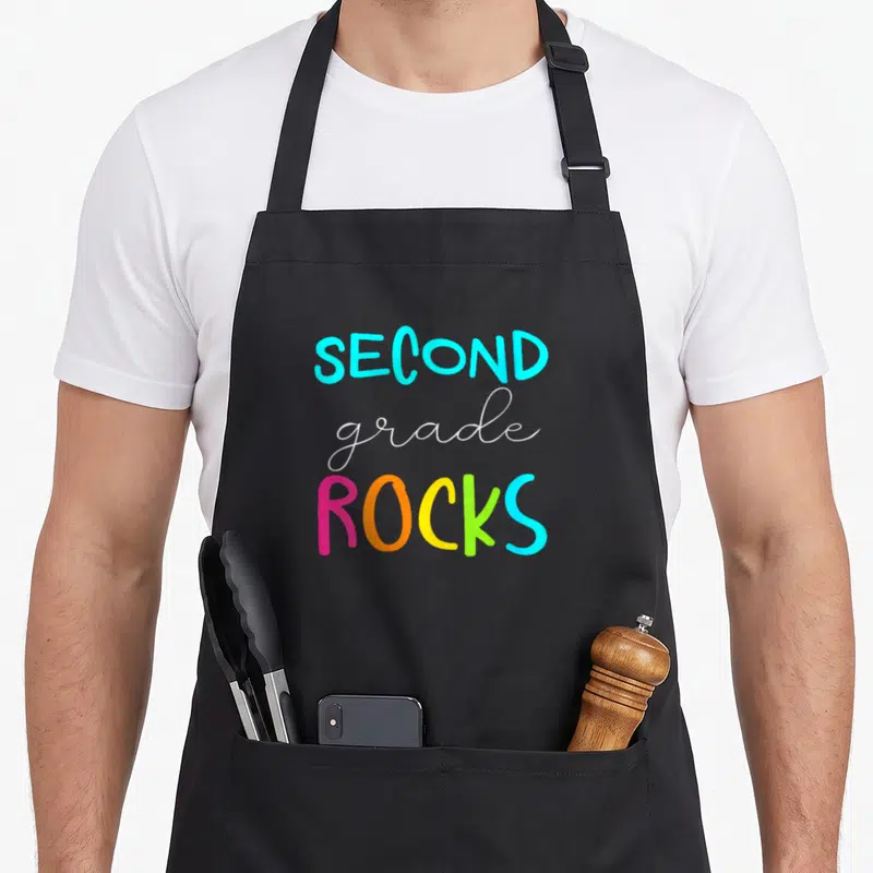 Rock 2nd apron - Stacked SECOND grade ROCKS text in blue white pink orange yellow and green typography — shown on a black cotton canvas apron against a white background