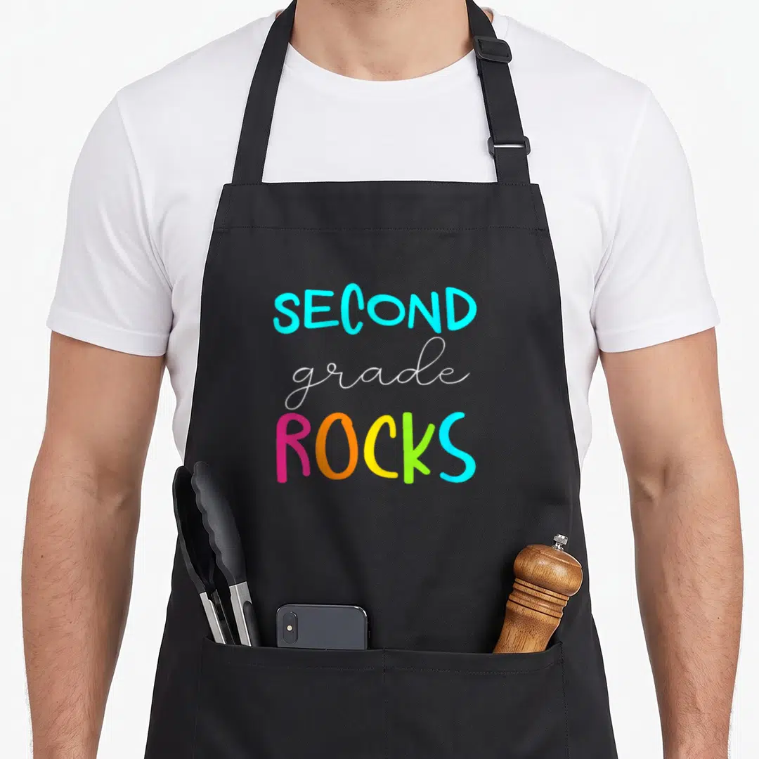 Rock 2nd apron - Stacked SECOND grade ROCKS text in blue white pink orange yellow and green typography — shown on a black cotton canvas apron against a white background