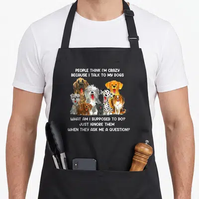 Cute talk cooking with dog apron - Nine cartoon dogs sandwiched between text about talking to pets on a black bib — shown on a black cotton canvas apron against a white background