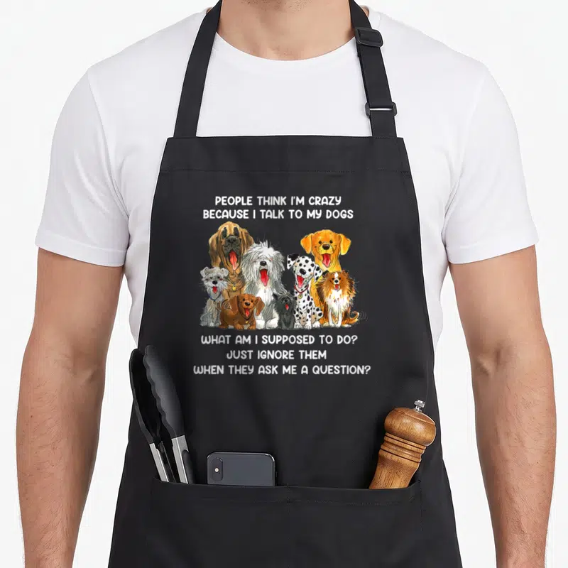 Cute talk cooking with dog apron - Nine cartoon dogs sandwiched between text about talking to pets on a black bib — shown on a black cotton canvas apron against a white background