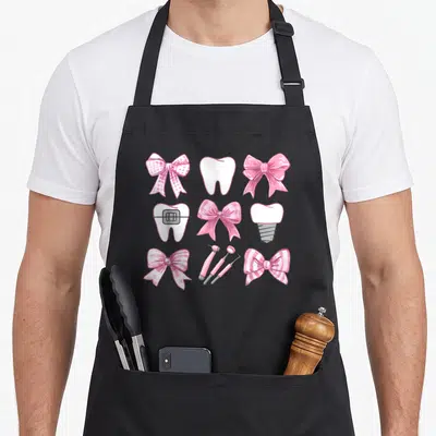 Coquette dentist apron - Pink bows, white teeth with braces, implant, and dental tools in a grid — shown on a black cotton canvas apron against a white background
