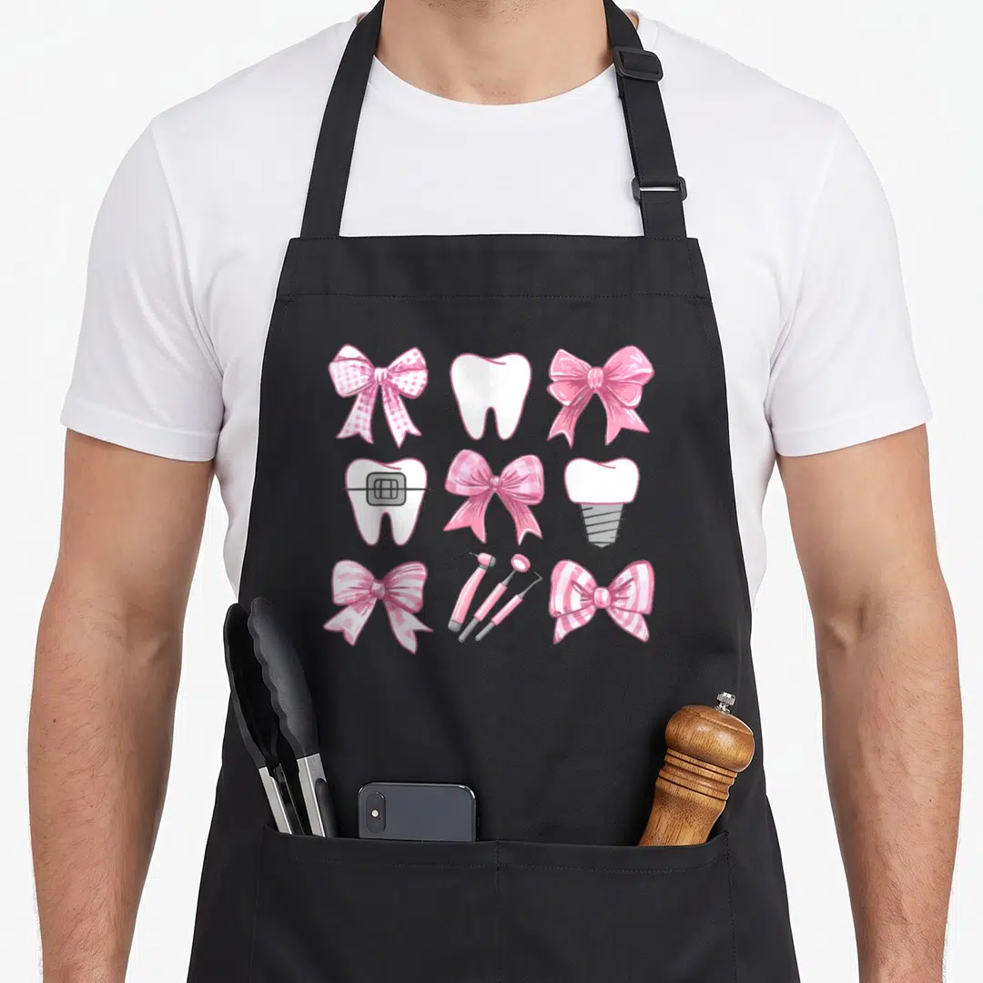 Coquette dentist apron - Pink bows, white teeth with braces, implant, and dental tools in a grid — shown on a black cotton canvas apron against a white background