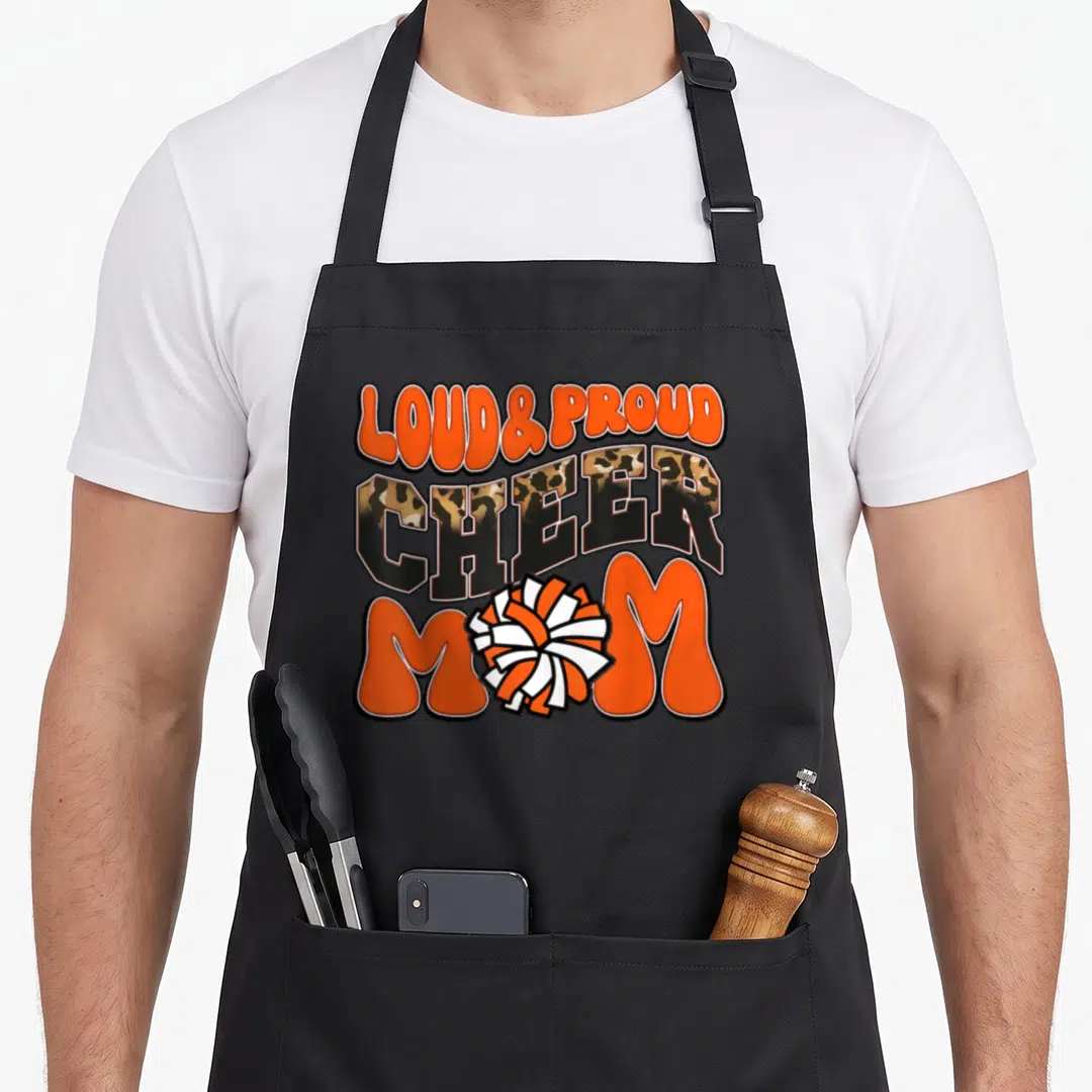 Proud crew aprons - Stacked LOUD PROUD CHEER MOM text with orange white pom-pom and leopard print CHEER letters — shown on a black cotton canvas apron against a white background