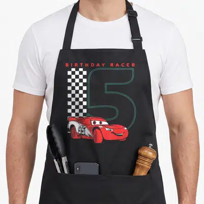 5th car apron - Red Lightning McQueen car overlapping a large grey number 5 next to a checkered flag with BIRTHDAY — shown on a black cotton canvas apron against a white background