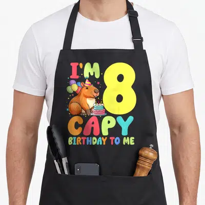 Capy 8th birthday apron - Cartoon capybara with party hat balloons and cake next to yellow 8 and colorful I'M 8 CAPY — shown on a black cotton canvas apron against a white background