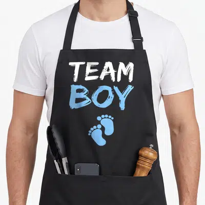 Shower gender reveal apron - Stacked TEAM BOY text with two light blue baby footprints on black fabric — shown on a black cotton canvas apron against a white background