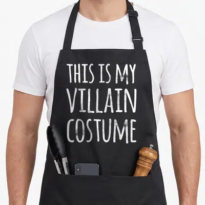 Villain funny halloween aprons - White stacked text reading THIS IS MY VILLAIN COSTUME on dark background — shown on a black cotton canvas apron against a white background