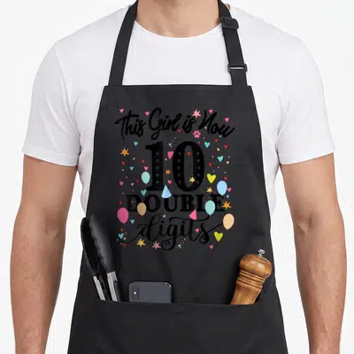 10th double apron - This Girl is Now 10 DOUBLE digits text with balloons hearts and stars — shown on a black cotton canvas apron against a white background
