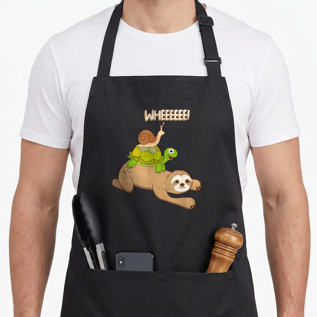 Decor sloth apron - Cartoon snail on turtle on sloth with WHEEEEEEE! text in brown and green — shown on a black cotton canvas apron against a white background
