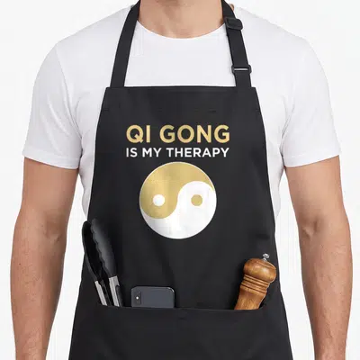 Gong chi qigong apron - Gold and white yin yang symbol below stacked text QI GONG IS MY THERAPY — shown on a black cotton canvas apron against a white background