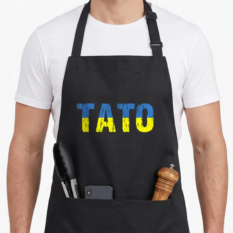 Distressed ukrainian aprons - TATO text split horizontally blue over yellow distressed font — shown on a black cotton canvas apron against a white background