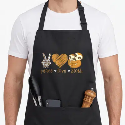 Peace sloth apron - Peace sign hand filled with tiny sloths, orange heart, and two hugging sloths with text — shown on a black cotton canvas apron against a white background