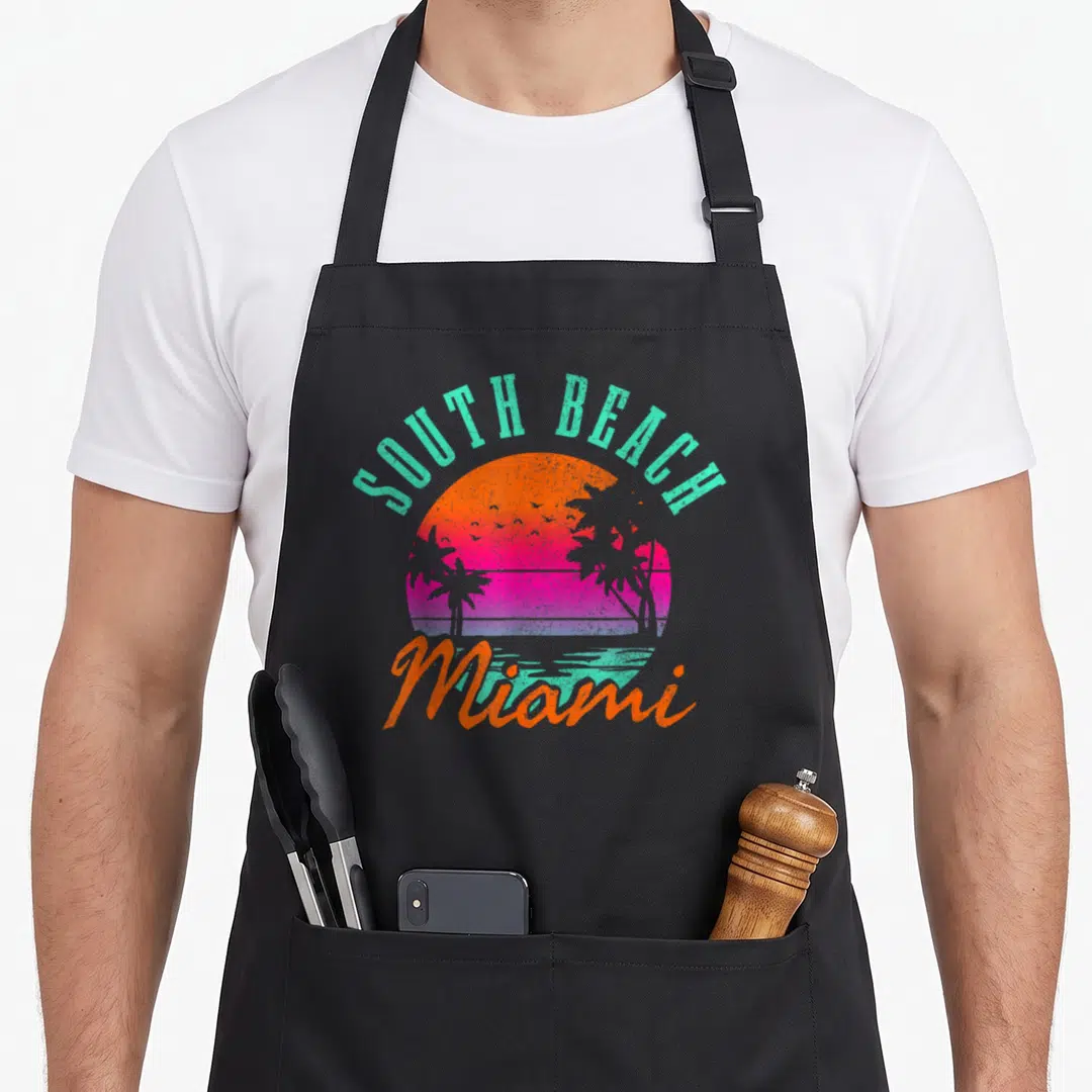 Beach miami florida apron - Retro sunset graphic with arched text and palm trees in teal orange pink purple — shown on a black cotton canvas apron against a white background