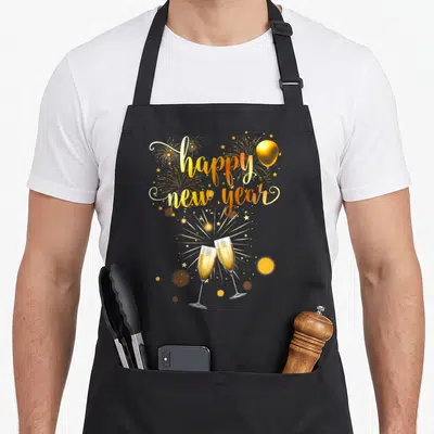 Supply christmas eve apron - Gold happy new year text with clinking champagne glasses and fireworks — shown on a black cotton canvas apron against a white background