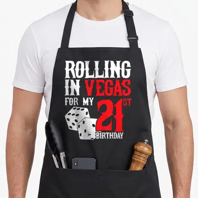 Vegas birthday apron - Rolling in Vegas 21st Birthday text with two dice and distressed red font — shown on a black cotton canvas apron against a white background
