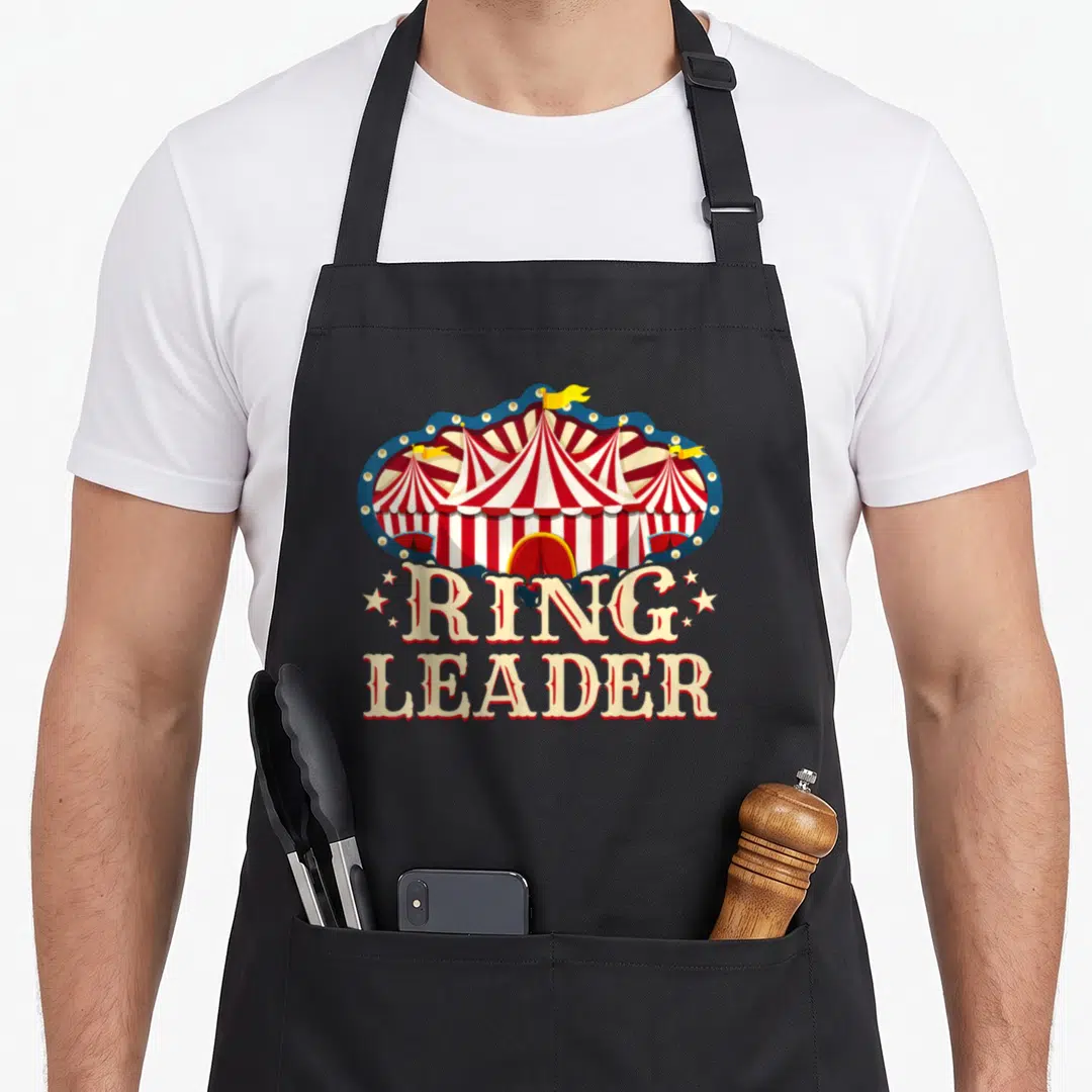 Ringleader birthday apron - Red white striped circus tent with blue scalloped border and stacked RING LEADER text — shown on a black cotton canvas apron against a white background