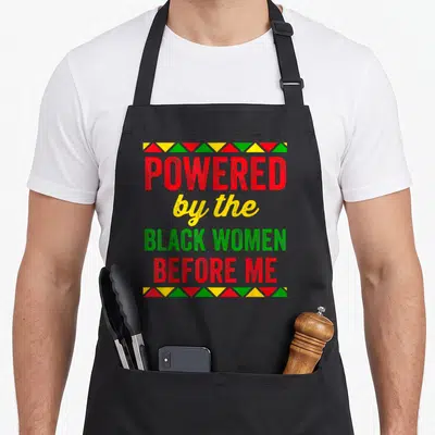 Powered mom daughter apron - Stacked POWERED by the BLACK WOMEN BEFORE ME text in red yellow green with triangle — shown on a black cotton canvas apron against a white background