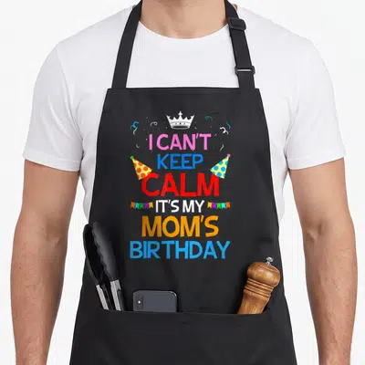 Funny nature birthday apron - Stacked colorful text I Can't Keep Calm It's My Mom's Birthday with crown and party hats — shown on a black cotton canvas apron against a white background